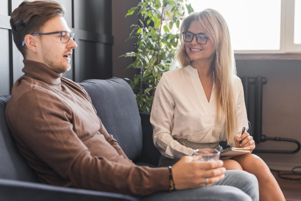 A man and a woman sitting on a couch talking. The woman is taking notes on a pad of paper.